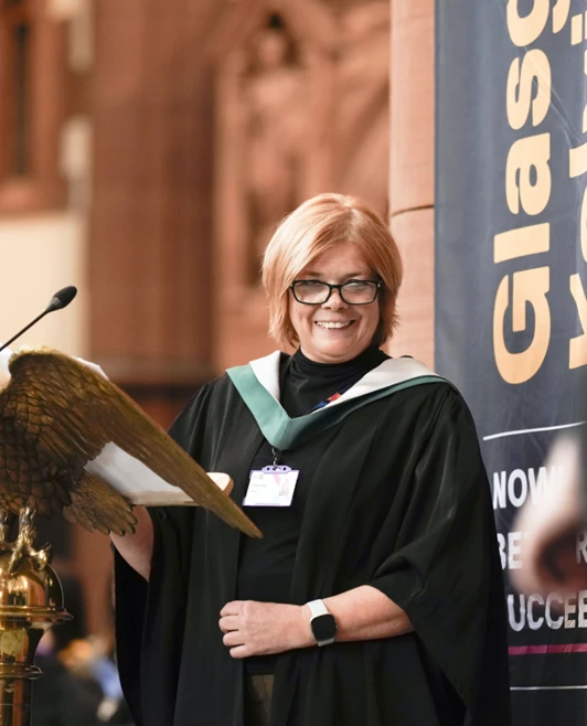 A smiling staff member at the podium during the Glasgow Kelvin College graduation ceremony, wearing a black gown with a green trim. A smiling staff member at the podium during the Glasgow Kelvin College graduation ceremony, wearing a black gown with a green trim.
